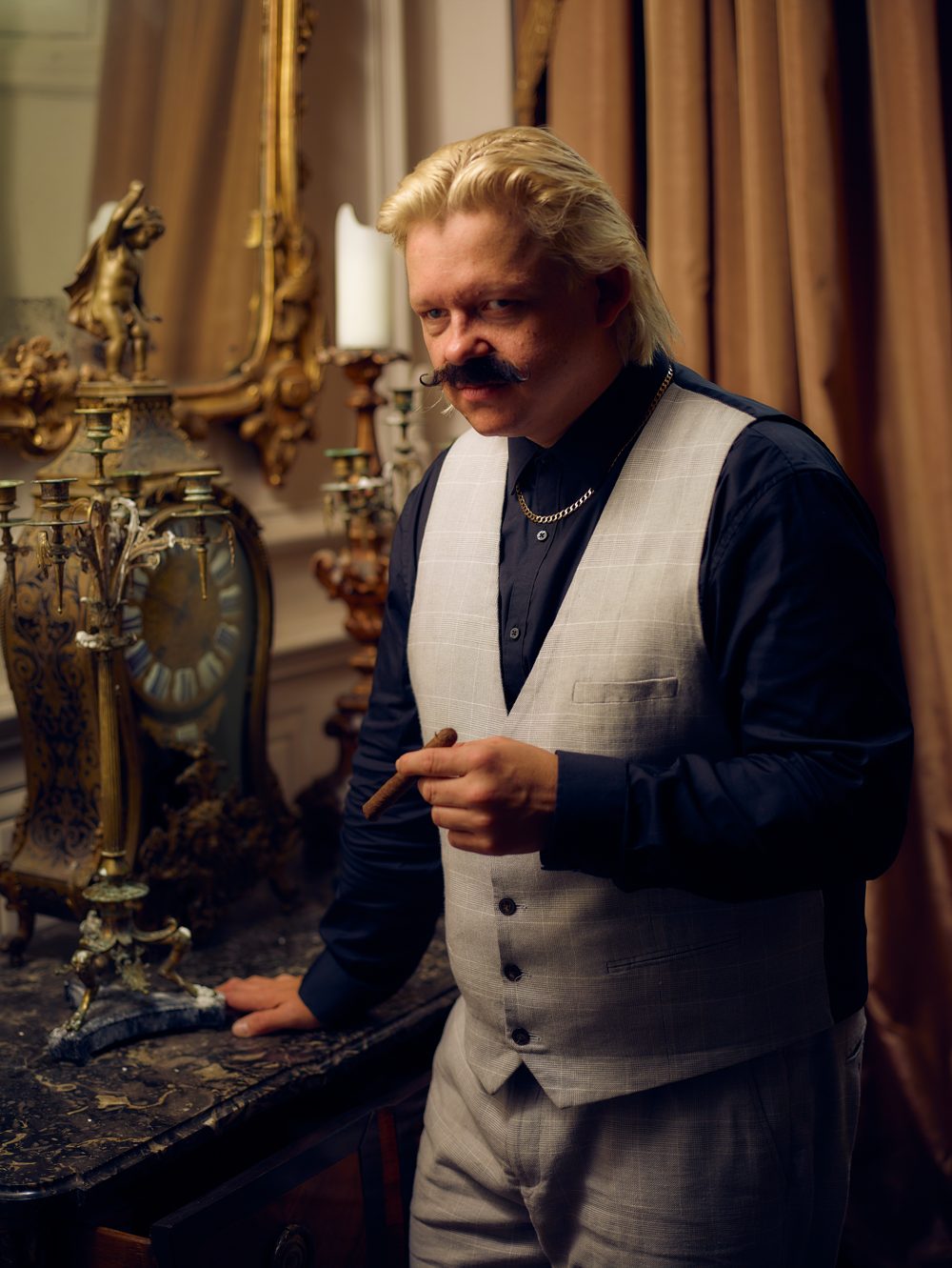 Jeremy Legal in a grey waistcoat holding a cigar beside a gilded mantel clock